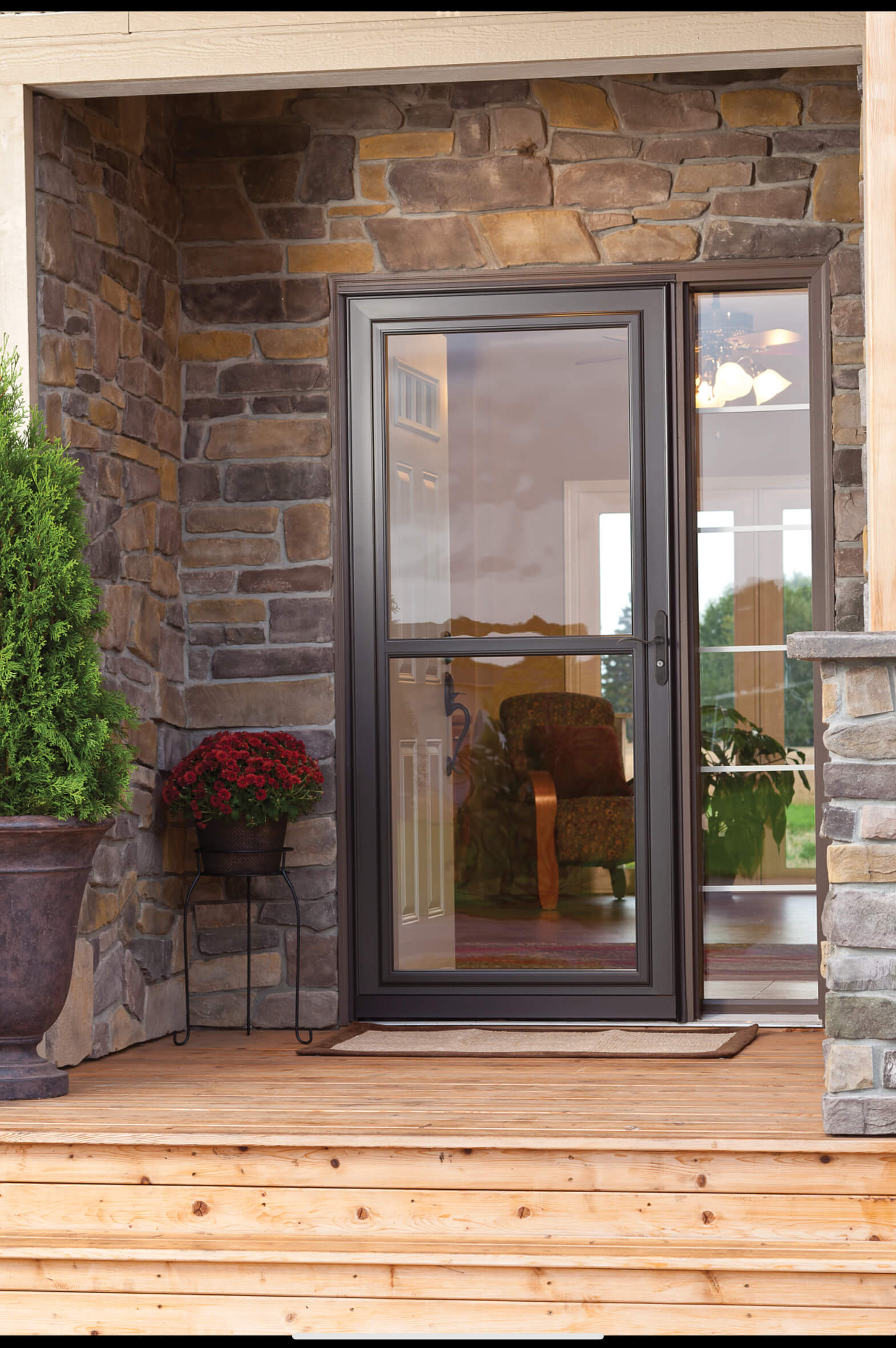 bronze-full-view-storm-door-stone-entryway Bronze full-view glass storm door installed on a stone entryway with a sidelight window and potted mums